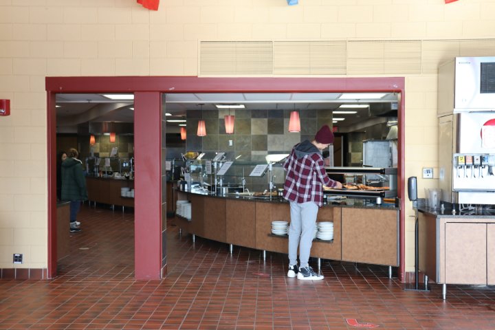 A student in a maroon beanie and plaid shirt stands in front of a food service counter, selecting items from a buffet. The dining hall features tile flooring and hanging pendant lights, with a beverage dispenser visible on the right.
